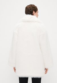 Cappotto in pelliccia sintetica bianca con colletto alto, vestibilità oversize e spalle scese. Texture liscia e design lungo fino al ginocchio. Nessuna chiusura visibile.