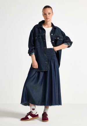 Denimjakke med store lommer, hvid t-shirt, lagdelt denimnederdel og burgundy sneakers med hvide detaljer. Strømperne har et mønster.