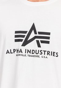 Tricou din bumbac alb, cu imprimeu grafic îndrăzneț în negru. Designul include litera stilizată "A" și textul "ALPHA INDUSTRIES".