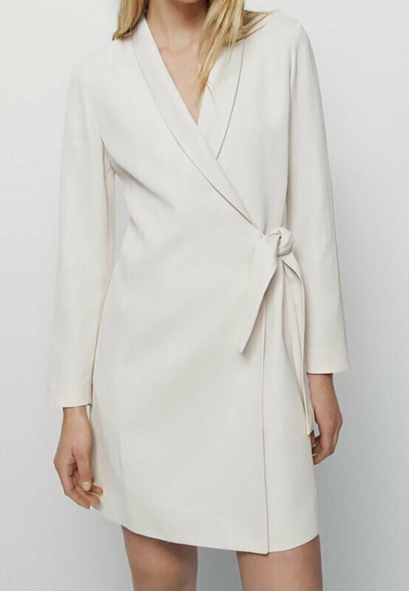 Femme portant une robe portefeuille blanche mi-longue à manches longues et ceinture nouée à la taille sur un fond uni.