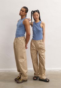 Two individuals stand back-to-back. They wear light blue ribbed tank tops, beige pleated trousers, and varied footwear.