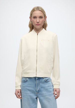 Femme blonde de face portant une veste crème zippée et un jean bleu clair sur fond blanc.