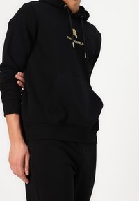 Felpa nera con logo dorato, dotata di tasca a marsupio frontale, cappuccio con cordino e polsini e orlo a coste. Texture liscia.