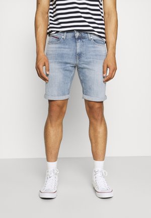 Homme portant un short en jean bleu clair retroussé, un t-shirt rayé noir et blanc, des chaussettes blanches et des baskets montantes blanches, debout devant un fond uni.