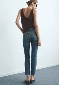 Brun ärmlös topp och blå jeans med medelhög midja och rak skärning. Jeans har bakfickor och en urtvättad finish. Modellen bär bruna klacksandaler.