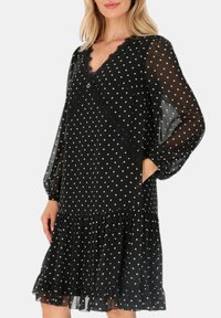 Czarna sukienka w kropki z długimi przezroczystymi rękawami, koronkowym wykończeniem, dekoltem w serek i warstwowym, falbaniastym dołem. Posiada boczne kieszenie i akcenty guzikowe.