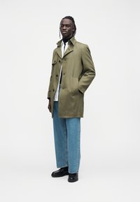 Trenci verde măsliniu cu două buzunare la piept, încheiat cu nasturi, purtat peste o cămașă albă cu guler și asortat cu blugi largi din denim și cizme negre.