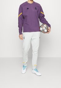 Sudadera morada con detalles amarillos, pantalones de chándal grises y botas de fútbol blancas. Sosteniendo un balón de fútbol con un patrón de estrellas. Logotipo del equipo visible.
