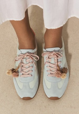 Feet wearing light blue sneakers with pink laces and decorative pom-poms, standing on beige carpet under a white skirt.