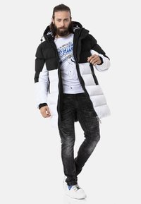 Schwarze und weiße Pufferjacke mit Kapuze, ausgestattet mit einem Reißverschluss und elastischen Bündchen. Getragen über einem grafischen weißen T-Shirt und dunklen Jeans.
