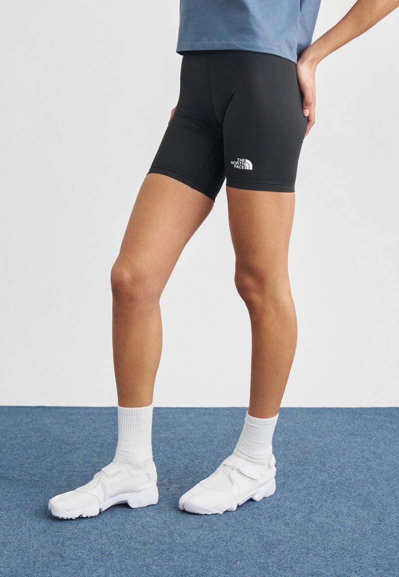 Persona che indossa pantaloncini da ciclismo neri The North Face, calzini bianchi e scarpe da ginnastica bianche, in piedi su un pavimento con moquette blu.