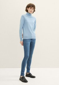 Suéter de cuello alto en azul claro, hecho de tejido de punto, combinado con jeans oscuros de corte ajustado y zapatos negros de cordones, contra un fondo neutro.