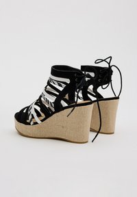 Las sandalias cuña en blanco y negro cuentan con correas entrelazadas de ante y un tacón de yute tejido. El diseño incluye cintas y una parte delantera descubierta.