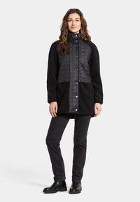 Musta tikattu takki, jossa on lampaantaljakäsivarret ja helma, korkea kaulus ja napillinen suljenta, yhdistettynä mustiin kapealahkeisiin farkkuihin ja saappaisiin.