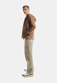 Joven de pie de lado con las manos en los bolsillos, vistiendo una chaqueta con capucha marrón, pantalones cargo beige y zapatos de senderismo blancos.