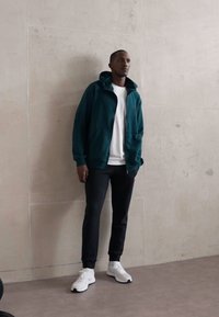 Dark teal zip-up hoodie with large front pockets, paired with a white T-shirt, black pants, and white sneakers. Smooth fabric and relaxed fit.