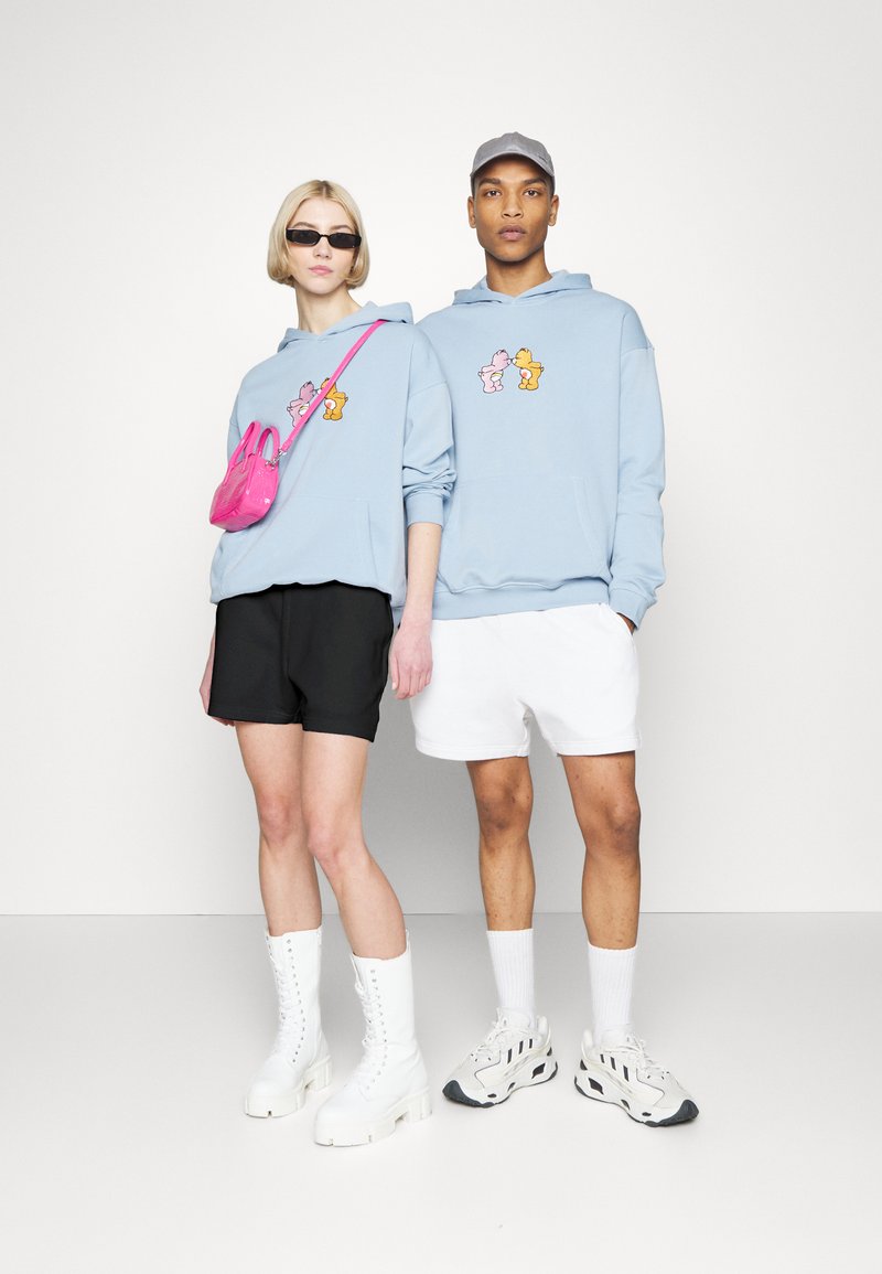 Lys blå hettegenser med tegneseriegraphikk, svarte shorts, hvite shorts, hvite snøreboots og en rosa håndveske. Moderne treningstøy.