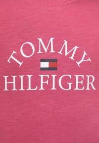 Sudadera rosa con texto en blanco que dice "TOMMY HILFIGER" y un logo rectangular en rojo, blanco y azul encima del nombre. Textura suave.