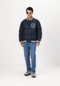 Námornícky puffer kabát s bielymi prúžkovanými manžetami a golierom, so zvýrazneným logom "C". Oblečené s modrými džínami a šedými športovými topánkami.