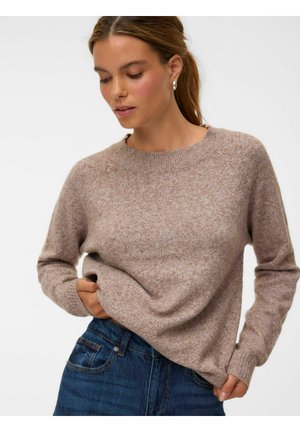 Femme aux cheveux châtain clair portant un pull en maille beige et un jean bleu, regardant vers le bas tout en ajustant son pull avec les mains.
