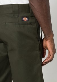 Pantalon de travail vert foncé avec une poche arrière et un patch logo Dickies, porté par une personne avec un bras et une main de teint foncé visibles.