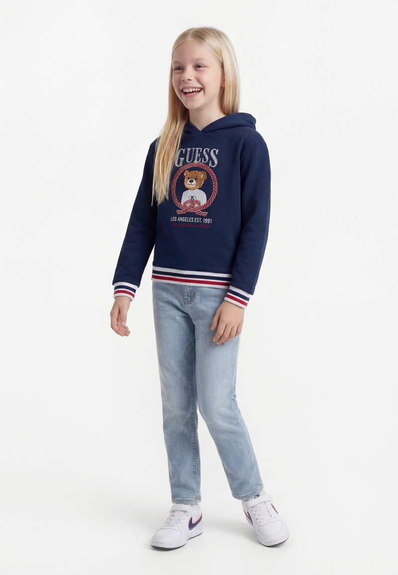 Κορίτσι που στέκεται και χαμογελά φορώντας μπλε σκουρόχρωμο φούτερ με κουκούλα και γραφικό αρκούδας, ανοιχτόχρωμο μπλε τζιν και λευκά αθλητικά παπούτσια σε μονόχρωμο φόντο.