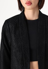 Femme portant un blazer en dentelle noire sur un crop top noir côtelé, avec des boucles d'oreilles créoles et des lèvres roses brillantes, sur fond blanc.