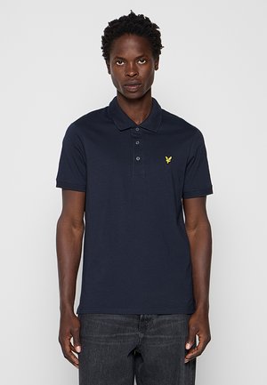 Mand iført en mørkeblå polo-shirt med en gul fuglelogo på venstre bryst, kombineret med mørke jeans, stående mod en enkel baggrund.
