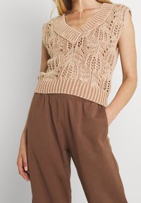 Beige stickad ärmlös topp med en djup V-ringning, texturerad spetsmönster och ribbad nederkant, ihopparad med bruna, löst sittande byxor.