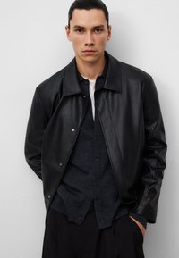Giacca in pelle nera con chiusura a scatto, texture liscia e una vestibilità leggermente sovradimensionata, indossata sopra una camicia button-up scura.
