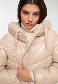 Giacca puffer beige con finitura lucida, caratterizzata da un colletto alto, cappuccio staccabile e cordoncini elastici. Chiusura con zip e motivo trapuntato.