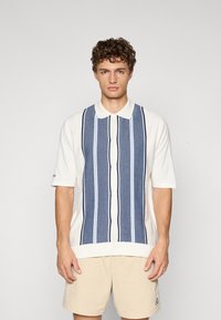 Camicia a maniche corte con corpo bianco, caratterizzata da righe verticali in blu navy e blu chiaro. Chiusura con bottoni e design del colletto classico.