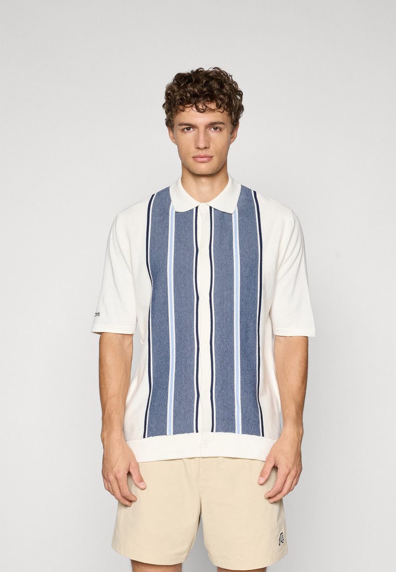 Camicia a maniche corte con corpo bianco, caratterizzata da righe verticali in blu navy e blu chiaro. Chiusura con bottoni e design del colletto classico.