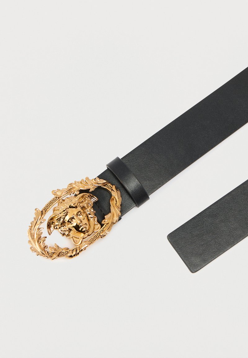 Svart läderbälte med ett utsmyckat, guldtonat ovalt spänne med ansiktsmotiv och blomdekorationer, kompletterat med ett texturerat band.