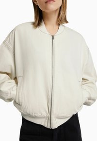 Svetle béžová bomber bunda vyrobená z měkké bavlny, s předním zipem, žebrovanými manžetami a bočn�ími kapsami. Má uvolněný střih a minimální švy.