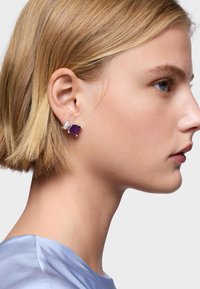 Orecchino a cuff in pietra preziosa viola, con taglio quadrato, impostato in argento. Modella con capelli biondi corti, indossa una maglia azzurra chiara. Vista in profilo.