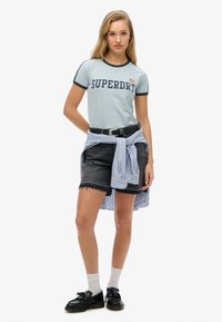 T-shirt bleu clair avec bordures navy et graphique "SUPERDRY", associé à un short en denim noir et une chemise à rayures bleu clair nouée autour de la taille.