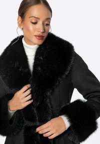 Černý kabát s velkým, měkkým límcem a manžetami z umělé kožešiny. Opasek v pase a vysoký límec doplňují elegantní design. Viditelná jemná struktura tkaniny.