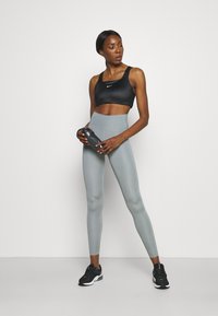 Tenue de sport composée d'un soutien-gorge de sport noir, de leggings taille haute gris clair et de sneakers noirs. Le modèle tient une bouteille d'eau noire.