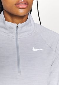 Gros plan d'une personne portant un haut zippé Nike gris clair avec un col montant et un logo Nike Swoosh blanc sur la poitrine.