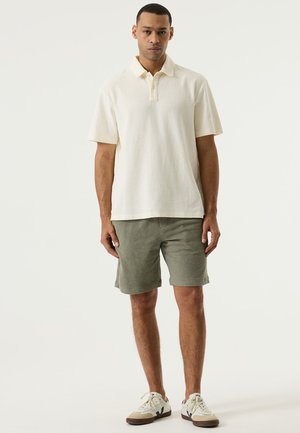 Roomwit polohemd met korte mouwen, textuurrijke stof, kraag met knoopsluiting. Olijfgroene korte broek met een zachte, geweven textuur. Sneakers in wit en beige.