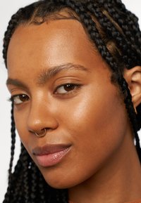 Primo piano di un volto con capelli neri intrecciati. Le caratteristiche includono una pelle liscia e un anello al setto nasale d'oro, con tonalità naturali in tutto.