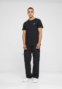 Black cotton t-shirt with a small white logo on the chest, paired with black cargo pants featuring multiple pockets and zippers, white sneakers.
