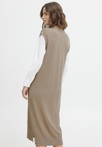 Abito senza maniche beige in maglia con silhouette dritta, spacchi laterali e una consistenza morbida; indossato sopra una camicia bianca con bottoni.