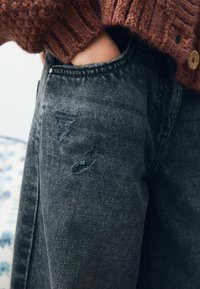 Musta farkut, joissa on kuluneita kohtia reiden yllä, yhdistettynä textured ruskeaan neulepaitaan, jossa on nappidetalji.