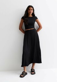 Top corto negro, falda larga plisada negra y sandalias negras con tiras. Tejido suave, corte holgado y esquema de colores neutros.
