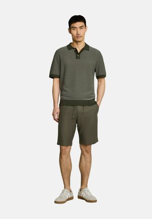 Man die een olijfgroen-wit gestreept poloshirt met korte mouwen draagt, olijfgroene shorts met trekkoord en witte sneakers, staand met één hand in de zak.