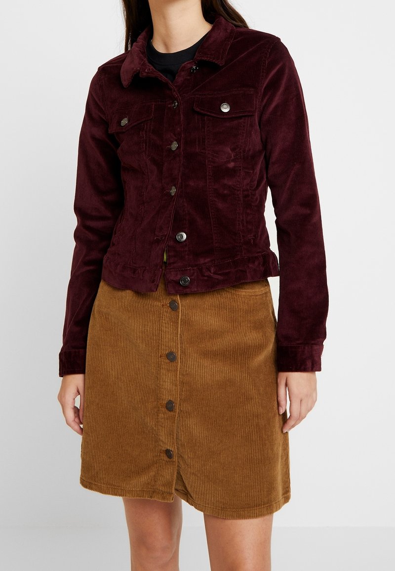 Femme portant une veste en velours côtelé bordeaux foncé avec des boutons métalliques et une jupe en velours côtelé marron boutonnée devant, debout sur un fond uni.