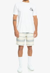 Bílá bavlněná tričko s černým logem na hrudi, zkombinované s béžovými pruhovanými kraťasy s světle modrými pruhy a bílými teniskami.