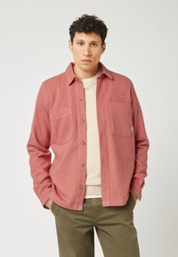 Man in een stofroze overhemd met knopen over een beige trui, in olijfgroene broek, staande met zijn handen in zijn zakken tegen een effen achtergrond.